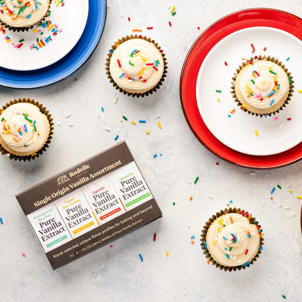 recipe image of vanilla cupcakes with vanilla buttercream and rainbow sprinkles. Five cupcakes are in the scene and two fo them are on blue, red, and white plates.. in the bottom left is Rodelle's single origin gift assortment