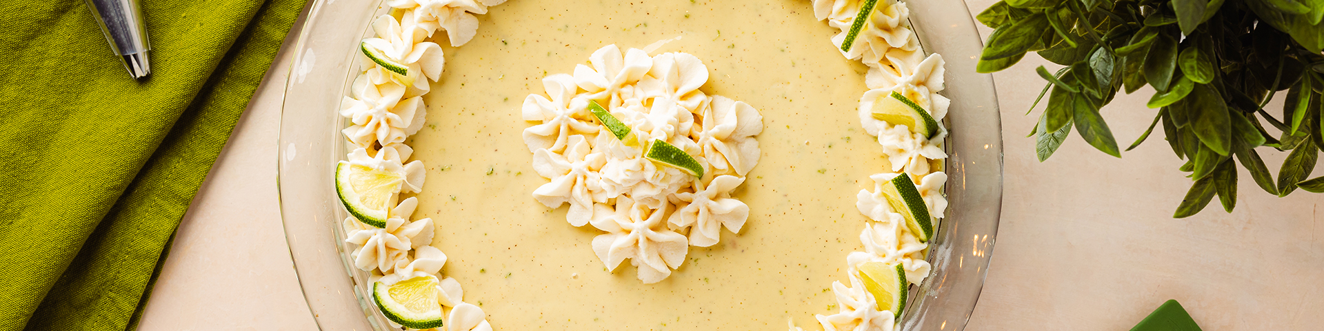 top view of a key lime pie with whipped cream, held by 2 hands and there are elements in the background: a piping bag of whipped cream, a green spatula and a plant