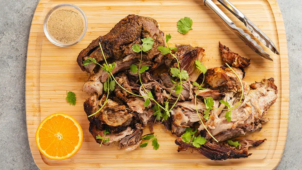 top view of a cuban bbq pork roast shredded on a wooden cutting board. On top of the pork roast is fresh cilantro. Tot he side of the pork roast is an orange cut in half, a pinch bowl with Rodelle's cuban seasoning, and a silver pair of tongs. The background is a neutral mid gray.