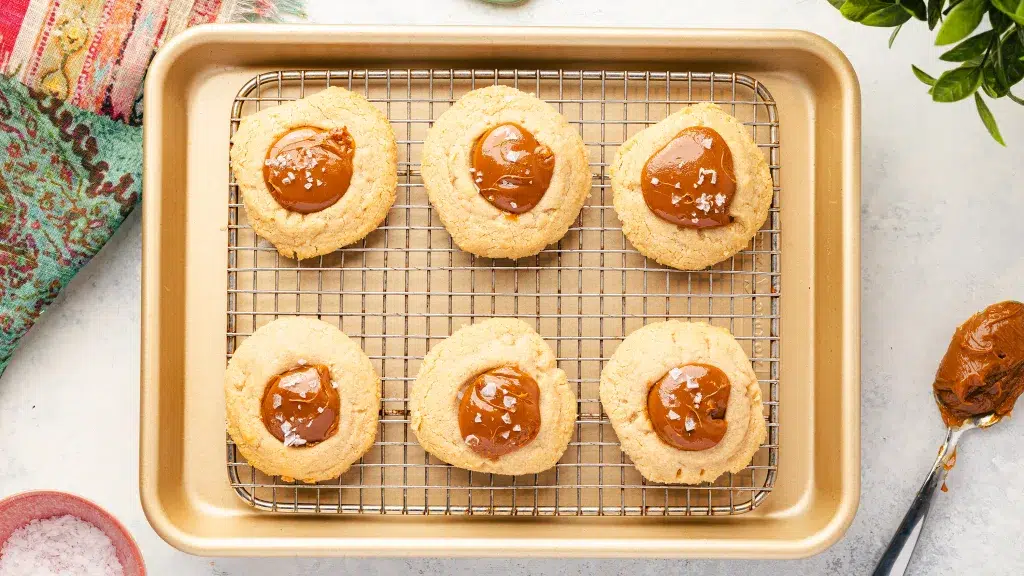 Top view of six salted dolce de leche cookies on a small brass pan with a cooling rack.Natural Baker's Extract. Surrounding the brass pan is a spoonful of homemade dolce de leche, a green, blue, and red towel, and a pinch bowl with salt.