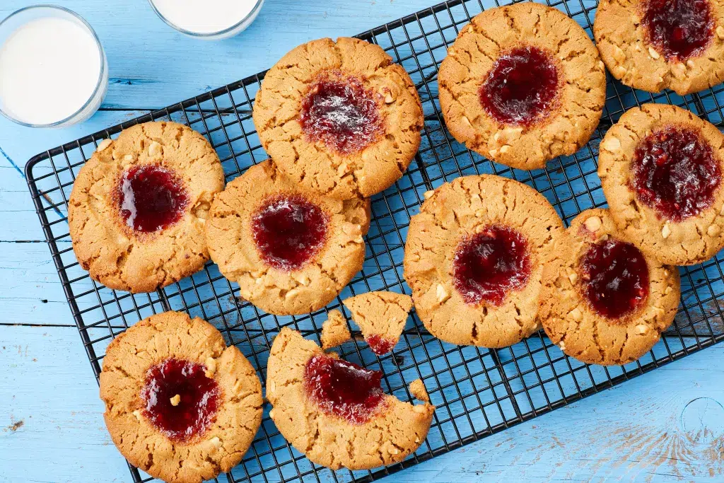 Peanut butter and jelly thumbprint cookies on cooling rack with a blue background and cup of milk