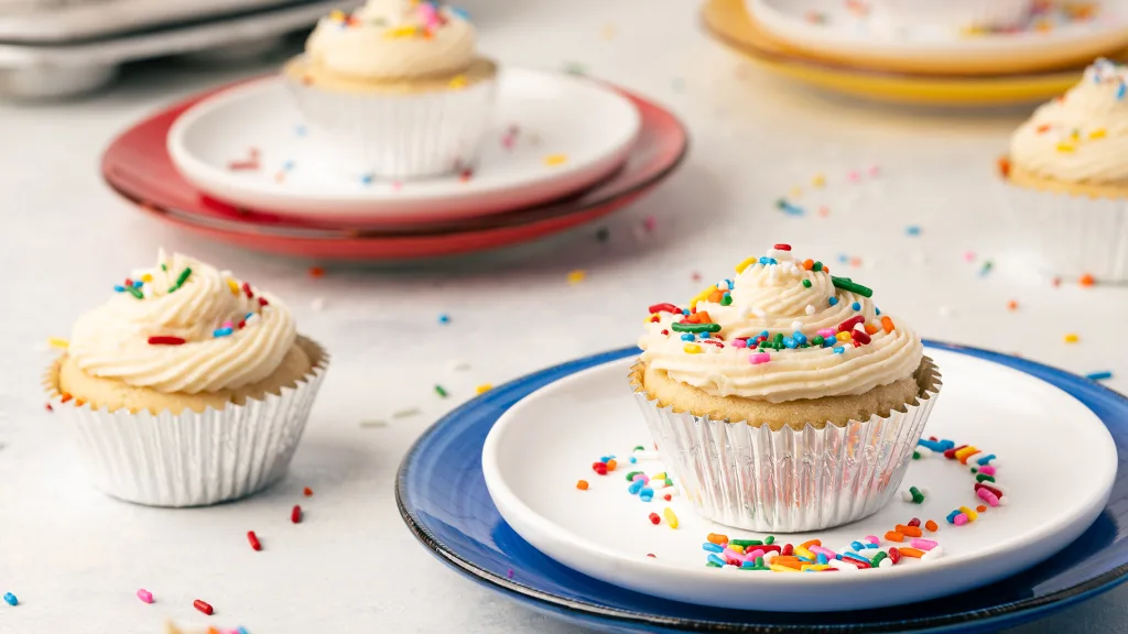side view of small batch vanilla cupcakes on a white and blue plate. There are additional cupcakes surrounding the scene, all in silver foil liners. Each cupcake is frosted with a vanilla buttercream and on top are rainbow sprinkles.