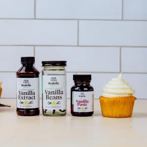 Side profile of a single double vanilla cupcake with vanilla bean frosting to the right. To the left is Rodelle's 2oz natural vanilla paste, 2 count gourmet vanilla beans, and Madagascar bourbon pure vanilla extract.