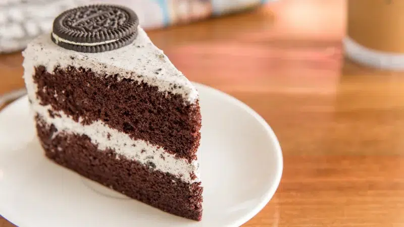 A slice of oreo cake showing two layers of chocolate cake and cream cheese frosting. On top of the slice is a single oreo. Cake stands on a white plate. Counter is a warm wooden oak color.