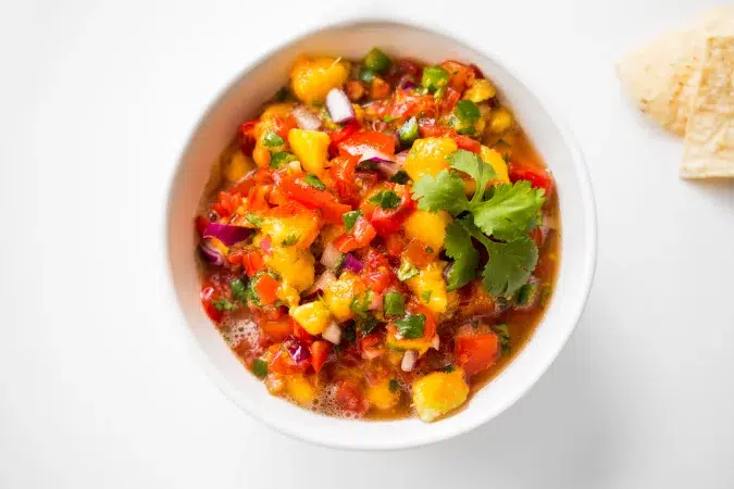 Above shot of a vanilla summer salsa in a white bowl. In the corner of the image are chips and the surface everything lays on is white.