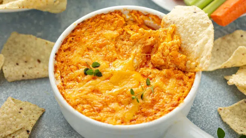 Close up of a white dipping bowl with handle containing Cuban Barbeque Chicken Cheese Dip. A round corn ship is dipped into the sauce showing the cheesy chicken dip. Surrounding the dip are more corn chips scattered throughout scene and in upper right hand corner are carrot and celery sticks.