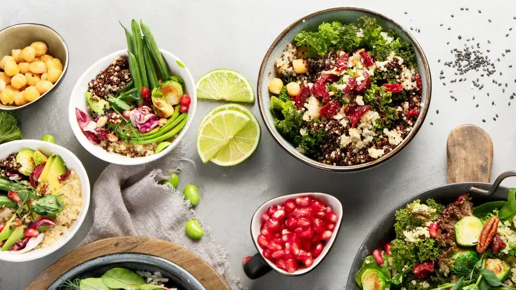 Above shot of Kale Salad with Quinoa and Pomegranate in a larger bowl. To side are smaller bowls of salad, slices of lime, pomegranates, and a tan dish towel. A family dinner set up.