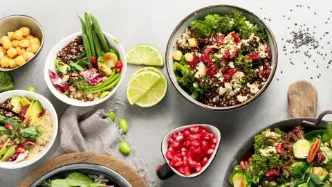 Above shot of Kale Salad with Quinoa and Pomegranate in a larger bowl. To side are smaller bowls of salad, slices of lime, pomegranates, and a tan dish towel. A family dinner set up.
