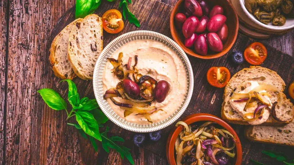 Whipped feta in a white bowl with grilled onions and tomatoes used for garnish on top. Surrounding the whipped feta is crusty bread, basil, tomatoes, grilled onions, and much more. Everything lays on a wooden table.