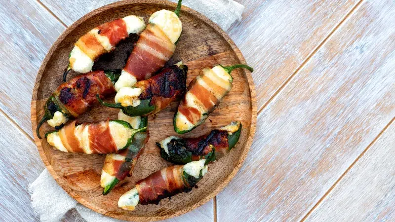 Top photo of cuban barbecue bacon wrapped jalapeño poppers on a large wooden plate. Underneath the plate is a white cloth and the background is an antique wooden white, light orange, and blue textured boards.