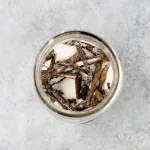 Top close up view of a mason jar containing dried chopped up vanilla beans in granulated white sugar, showing flecks of vanilla seeds spread throughout the sugar. The background is a light gray.