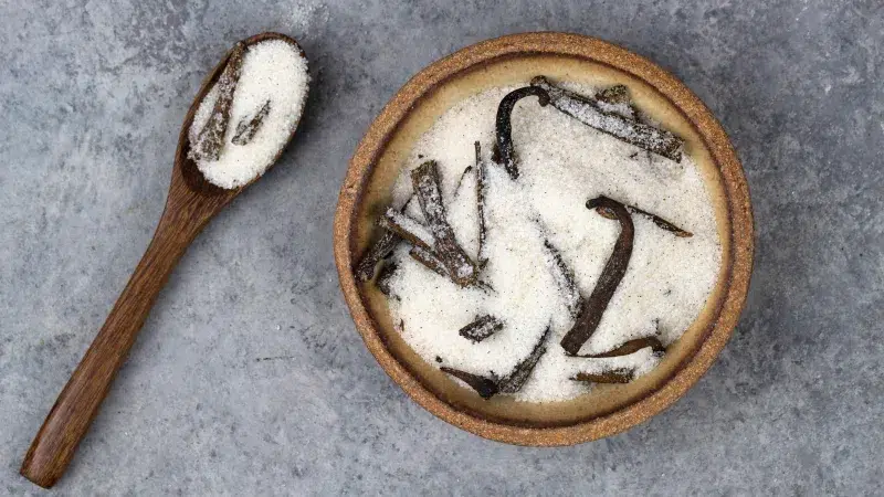 Top shot of granulated white sugar in a small earthtoned bowl. You can see flecks of vanilla seeds throughout the sugar along with dried out vanilla beans. To the left of the earth toned bowl is a dark brown wooden spoon with vanilla sugar. The background is a dark gray.