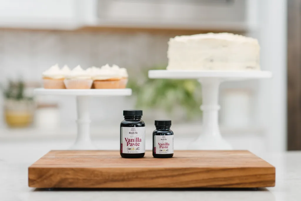 two bottles of vanilla paste on cutting board in front of a cake and cupcakes