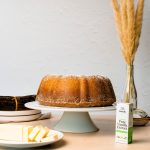 side photo of the kentucky butter bundt cake on a white cake stand. The foreground contains Rodelle's 2oz Pure Tanzanian Vanilla Extract and a plate with butter sliced up. the background contains a stack of speckled earth toned plates and a bundle of vanilla beans and a dried plant accessory like wheat. The background is white and the table is a tan and pink color. The image feels warm.