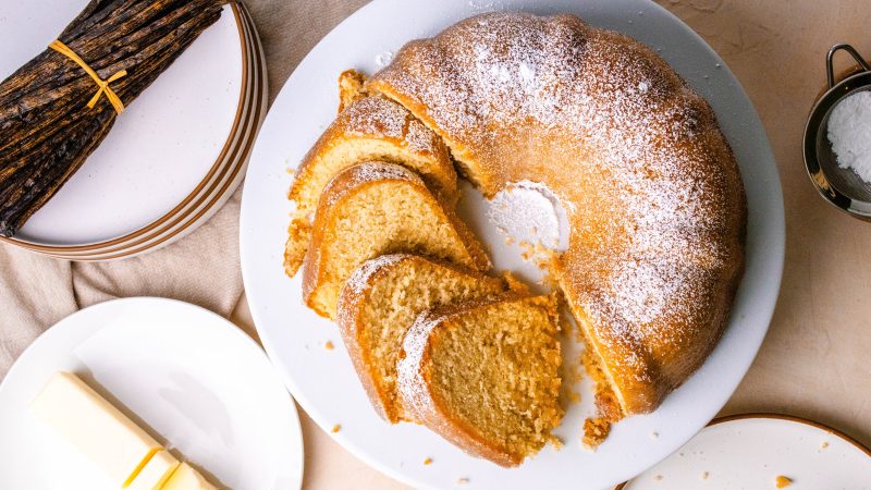top view of a kentucky butter bundt cake on a white cake stand. The cake is dusted with powdered sugar and a portion of it is sliced and laying sideways. Surrounding the bake is a small plate with cut up butter, a bundle of vanilla beans, plates, and a sifter with powdered sugar. The table is a light pink and tan color.
