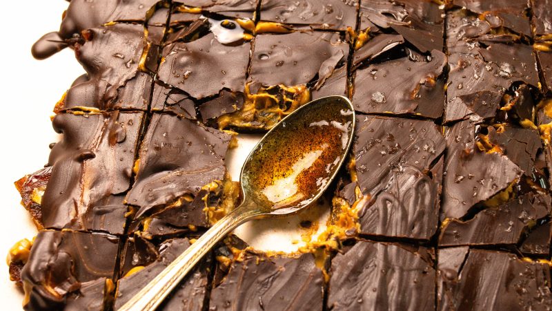 close up of vanilla peanut butter chocolate bark cut in slices. One slice has been taken out with a brass spoon replacing it containing vanilla paste where you can see the flecks of vanilla seeds.