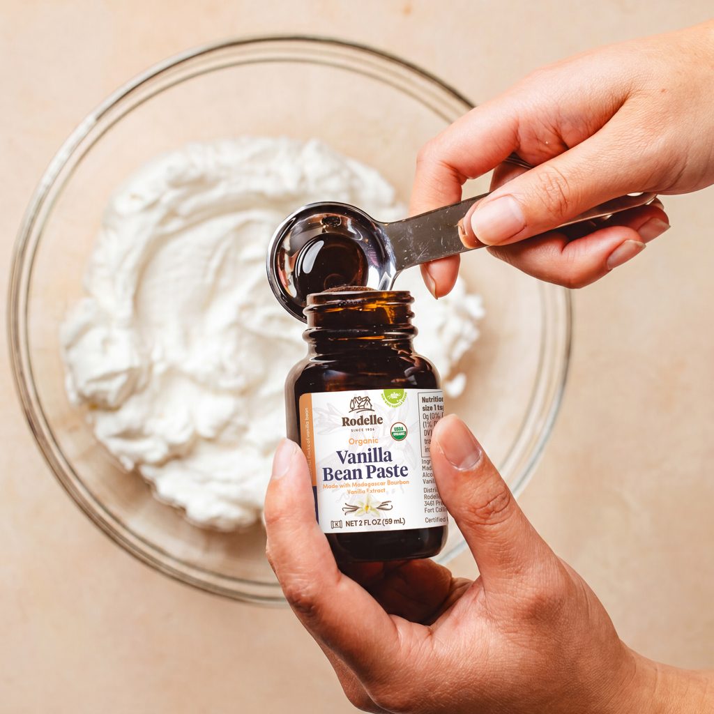 lifestyle image of hands pouring 2oz organic vanilla bean paste into a tablespoon. Below the vanilla paste and hands is a clear bowl is whipped cream. the table is a muted peach color