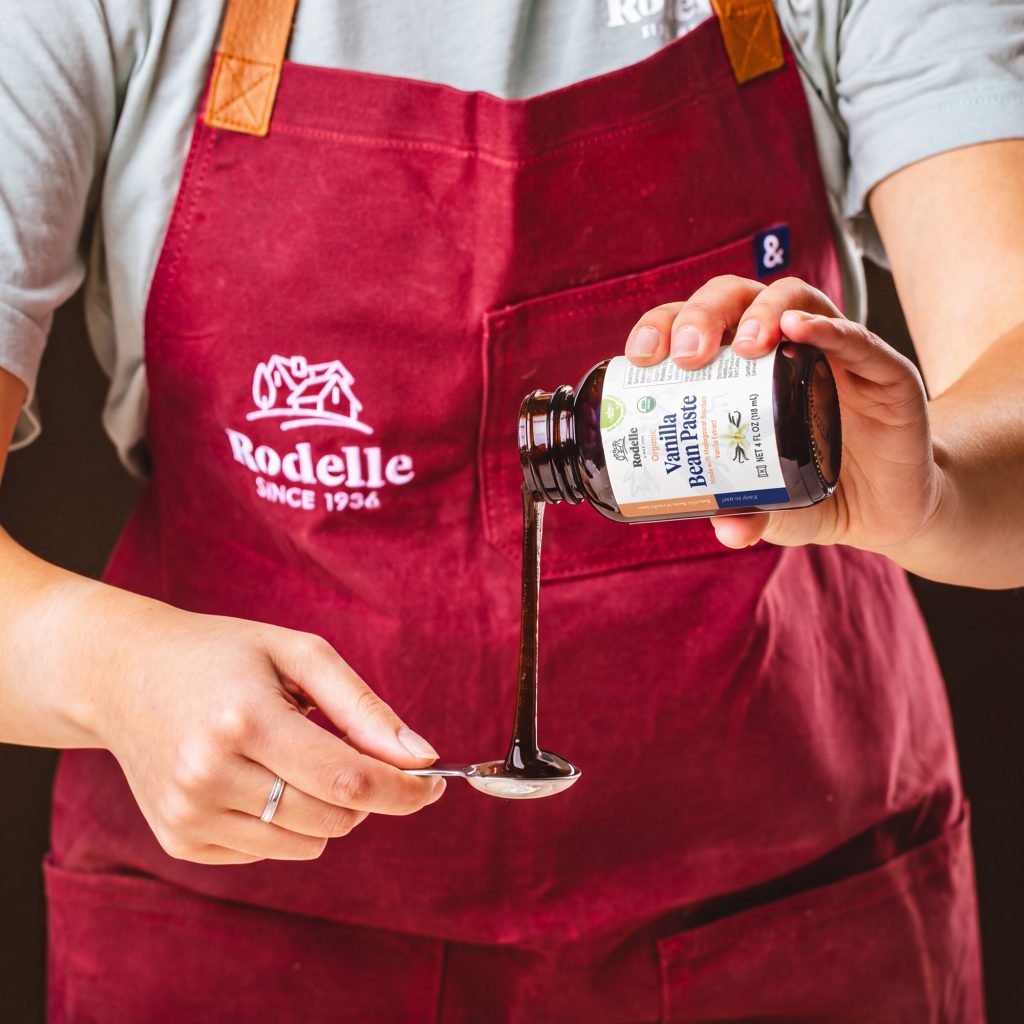 lifestyle image of a person pouring Rodelle's 4oz organic vanilla bean paste into a measuring spoon. the person is wearing a red apron with a white Rodelle logo