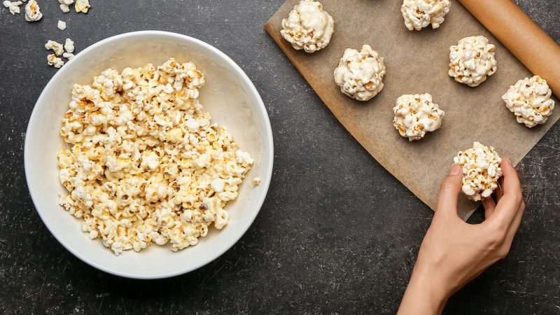 top photo of browned butter popcorn balls on a brown parchment paper in the upper right with a hand extending to grab one. To the left is a plate with additional popcorn.