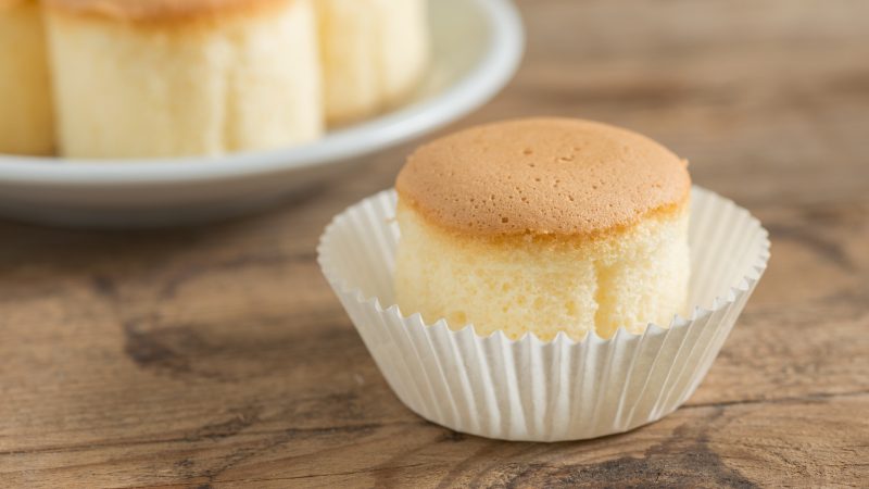 close up of a gluten free chiffon cupcake with a white wrapper slightly undone. The table is oak and in the upper left is a white plate with additional cupcakes.