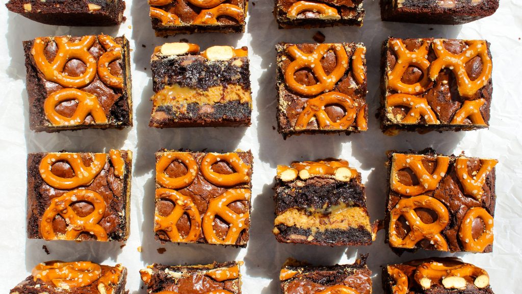 top photo of squares of midnight snack brownies organized in rows. two of the bars are flipped the opposite way so you can see four layers of brownie, cookie, and pretzels. all the bars have pretzels added. bars lay on parchment paper with sunlight entering from the upper portion of the photo casting long shadows downwards.