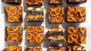 top photo of squares of midnight snack brownies organized in rows. two of the bars are flipped the opposite way so you can see four layers of brownie, cookie, and pretzels. all the bars have pretzels added. bars lay on parchment paper with sunlight entering from the upper portion of the photo casting long shadows downwards.