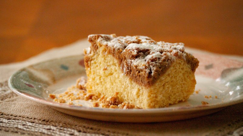 Close up of a slice of tanzanian vanilla crumb cake on a vintage plate and laced mat. The background is wooden. The crumb cake is cut in a rectangle where you can see a delicious cake and a nice layer of crumb topping and powdered sugar