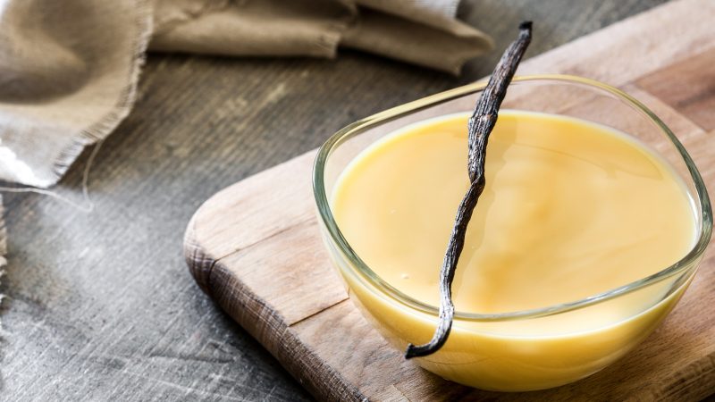 close up angle shot of a medium clear glass bowl containing creamy vanilla custard with a vanilla bean laying on top horizontally. The bowl lays on a wooden cutting board. The background is a dark gray texture and there is a light gray-ish tan cloth in the upper left.