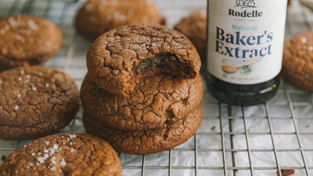 image of beautiful brownie cookies scattered on a cooling rack. Towards the middle are three cookies stacked o one another. You can see the flaky salt on top of each cookie. In the upper right is Rodelle's 4oz Natural Bakers Extract.