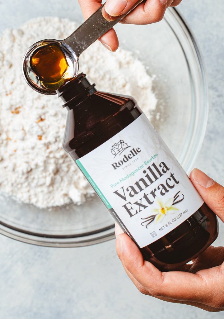 hand pouring 8oz rodelle madagascar bourbon pure vanilla extract into a measuring spoon. below the bottle being poured is a clear bowl with flour. the table is a light grayish blue color.