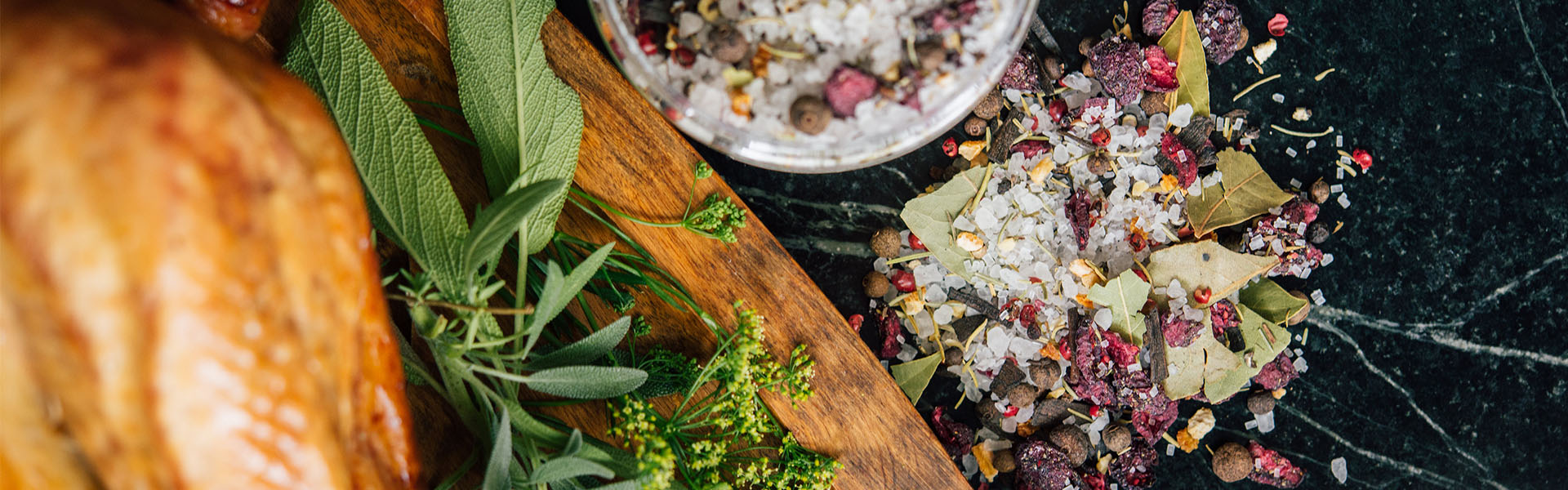 top image of a turkey with herbs next to it. There lays a Rodelle 13oz turkeyb rine seasonig container, opened. Next to it is a pile of turkey brine seasoning - you can see the salt, cranberries, and additional herbs and spices mixed together. The counter is a green gold color.